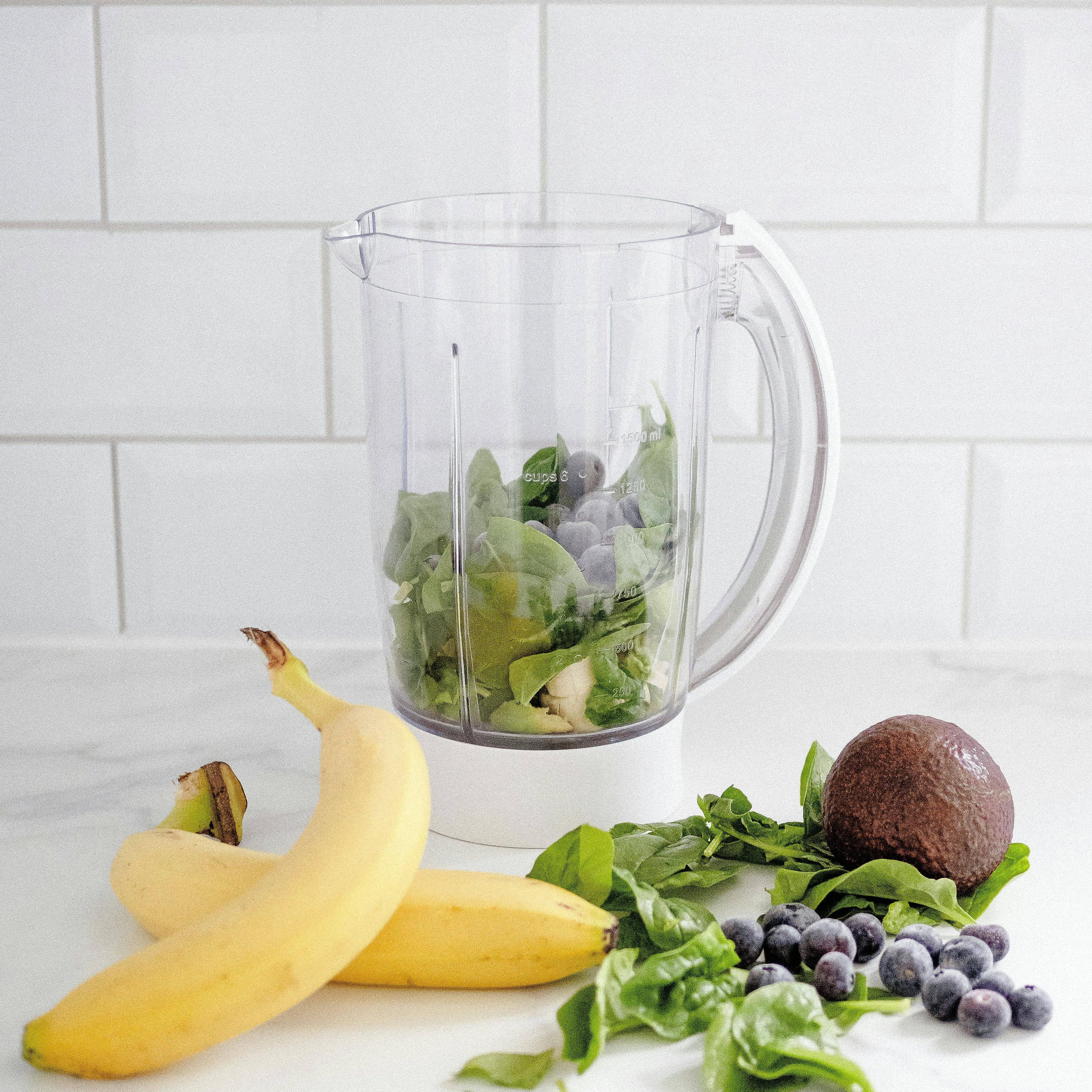 Healthy breakfast to balance blood sugar. Ingredients for a green smoothie, including bananas, spinach, avocado, and blueberries, arranged next to a blender jar on a white kitchen counter with a tiled backsplash.