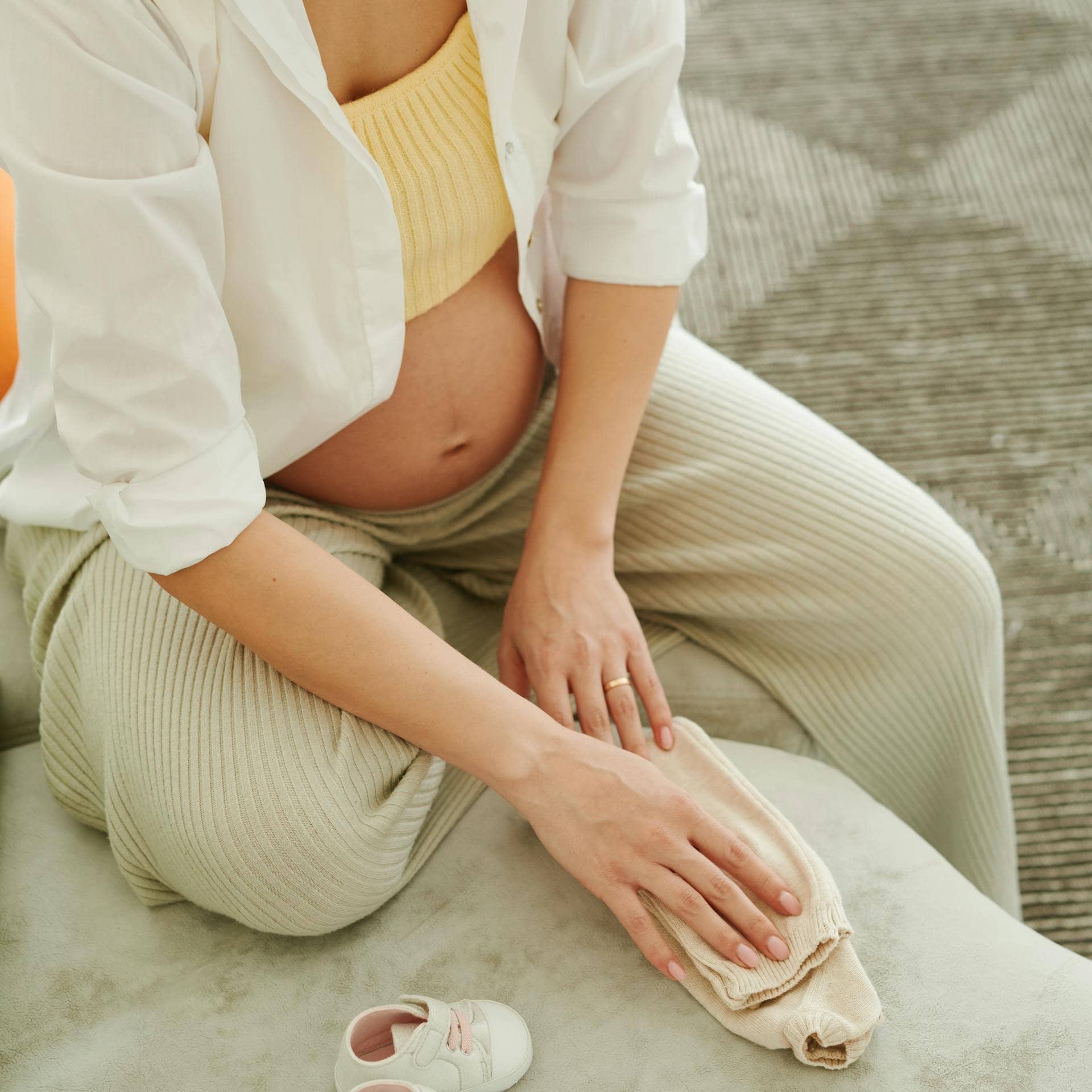 Pregnant woman during perimenopause folding baby clothes beside baby shoes.