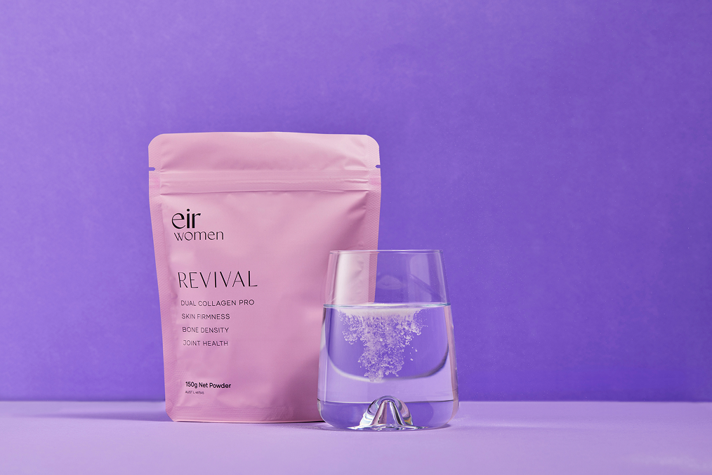 A pink pouch of Eir Women’s Revival Dual Collagen Pro sits next to a glass of water with collagen powder dissolving inside, set against a purple background.
