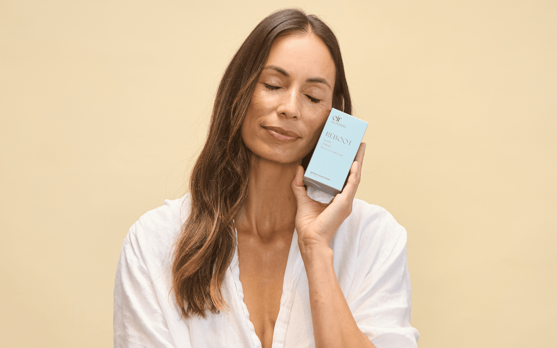 Woman with long brown hair in a white blouse holding Eir Women Reboot box to her face against a beige background