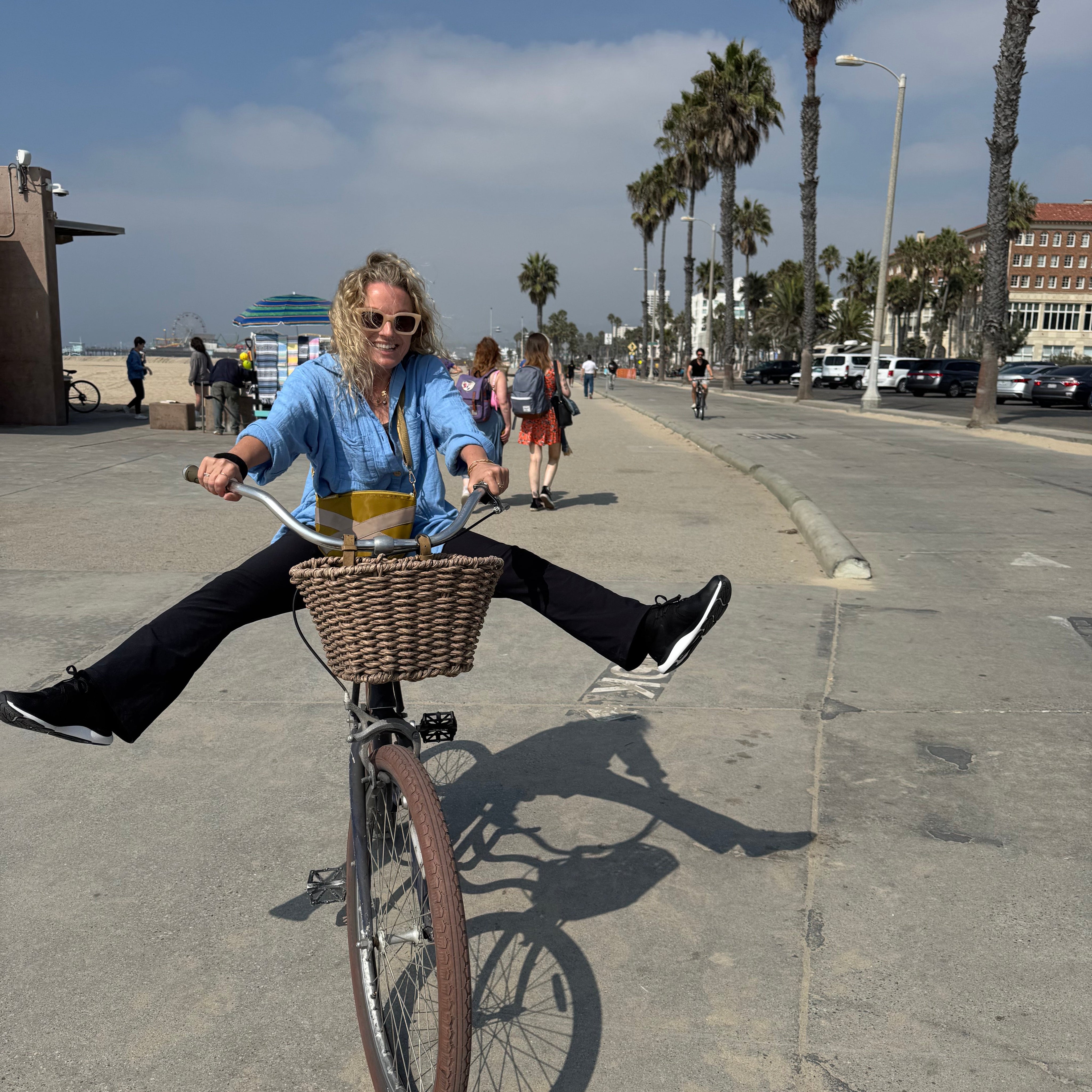 Lisa Walker, Co-Founder of Eir Women, riding a bike along the beachfront promenade in Los Angeles, smiling with legs outstretched on a sunny day.