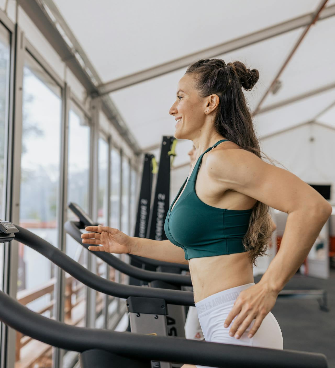 Woman running on a treadmill in a gym, wearing activewear and smiling while exercising near large windows.