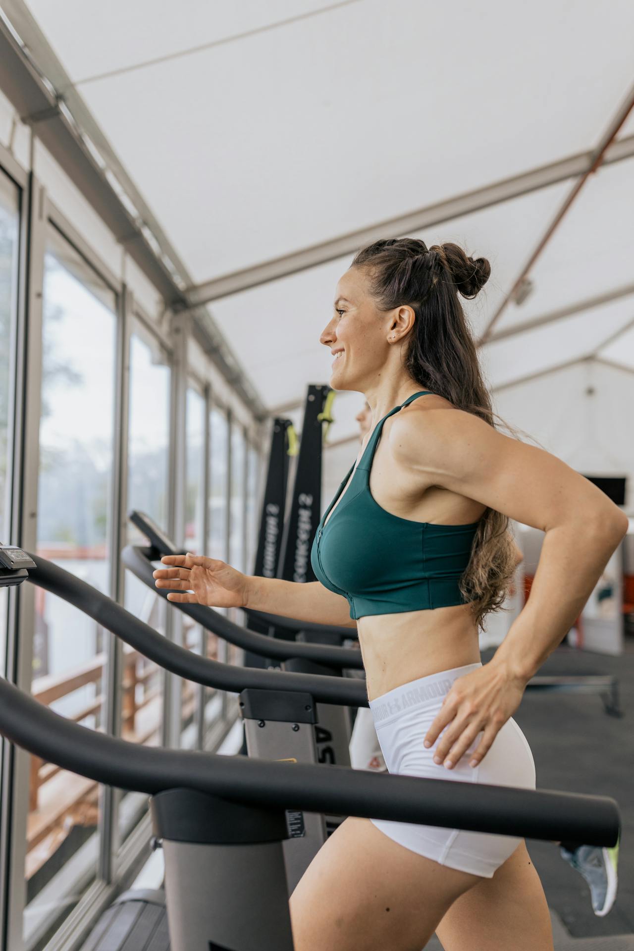 Woman running on a treadmill in a gym, wearing activewear and smiling while exercising near large windows.