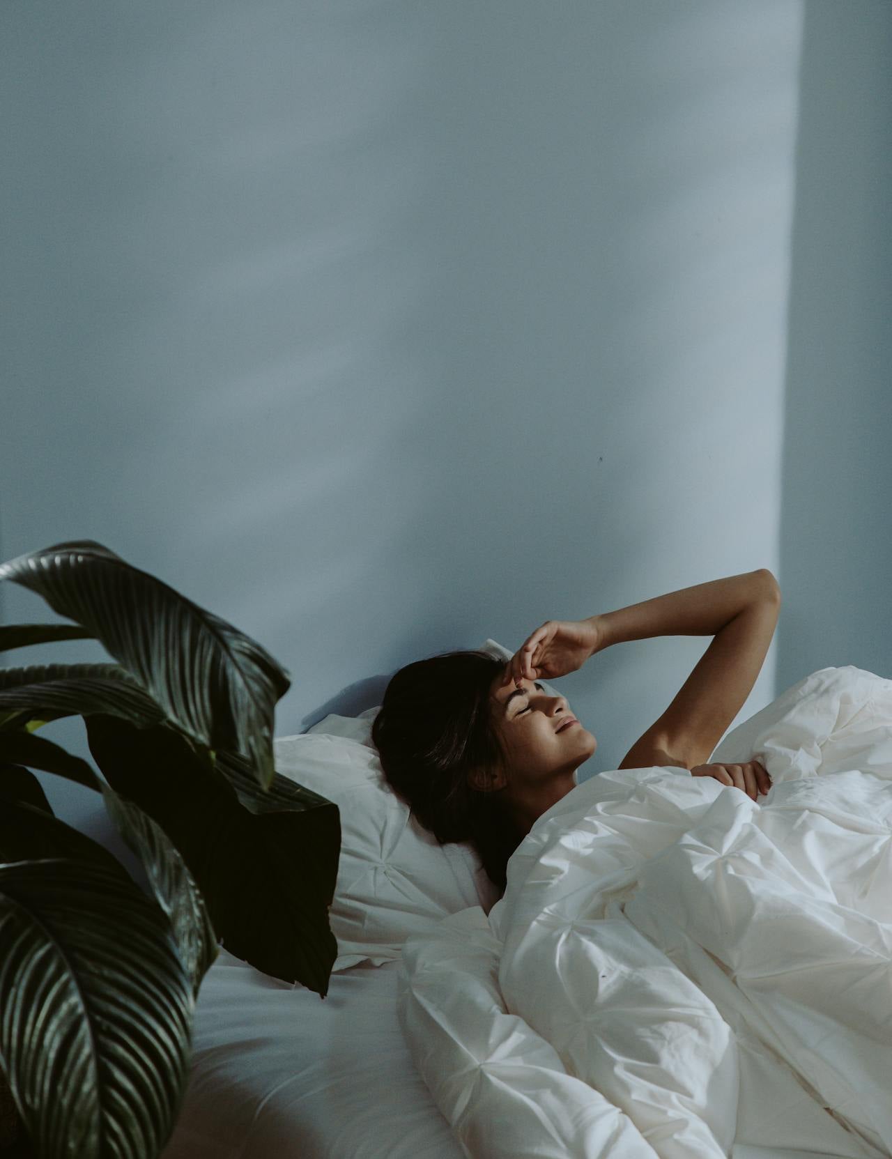 Woman lying in bed under white bedding, eyes closed with hand resting on her forehead, in a softly lit minimalist bedroom with a plant nearby.