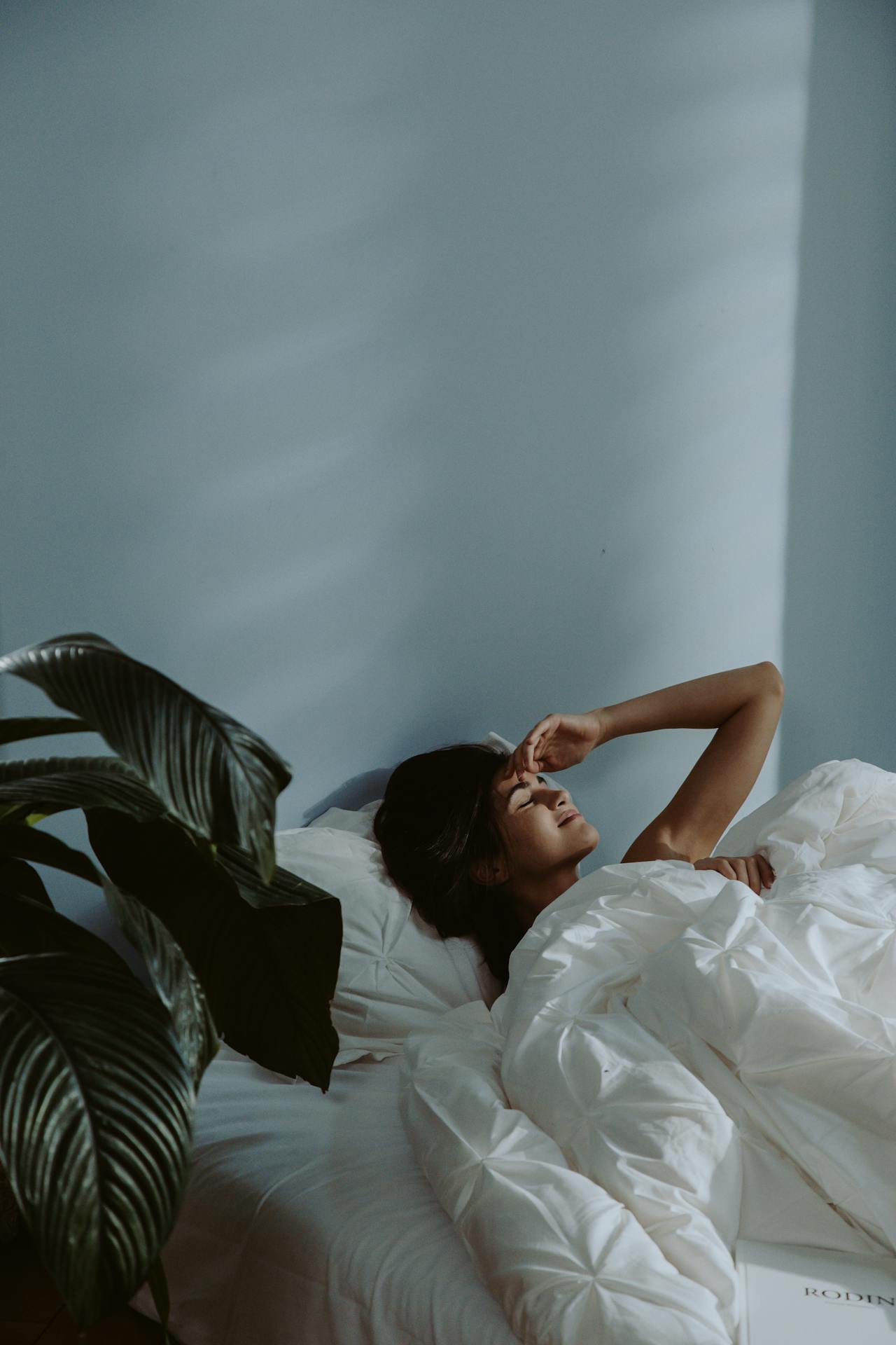 Woman lying in bed under white bedding, eyes closed with hand resting on her forehead, in a softly lit minimalist bedroom with a plant nearby.