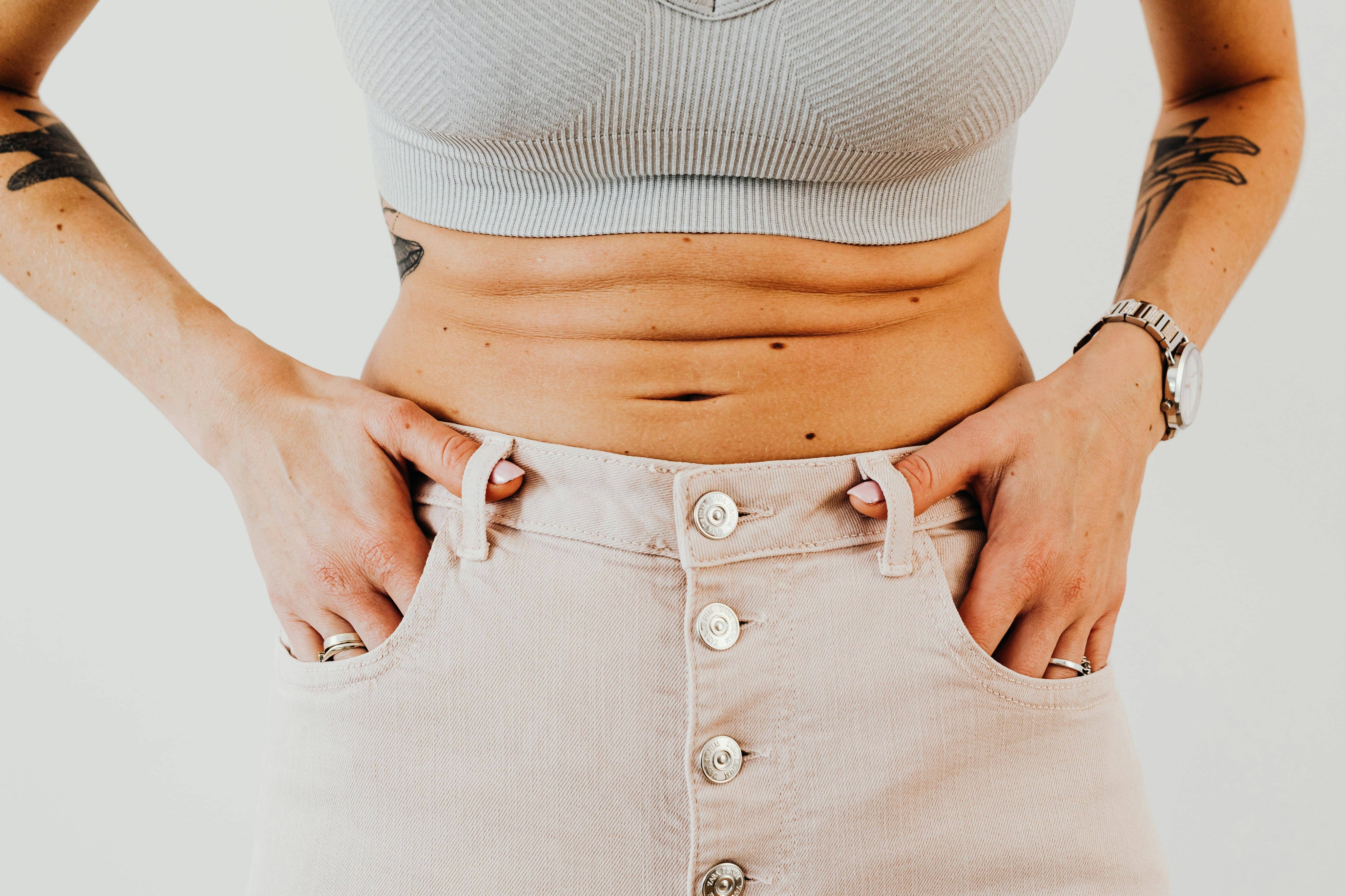 Close-up of a woman's midsection wearing high-waisted beige jeans and a ribbed crop top, showing natural belly folds and hands in pockets.