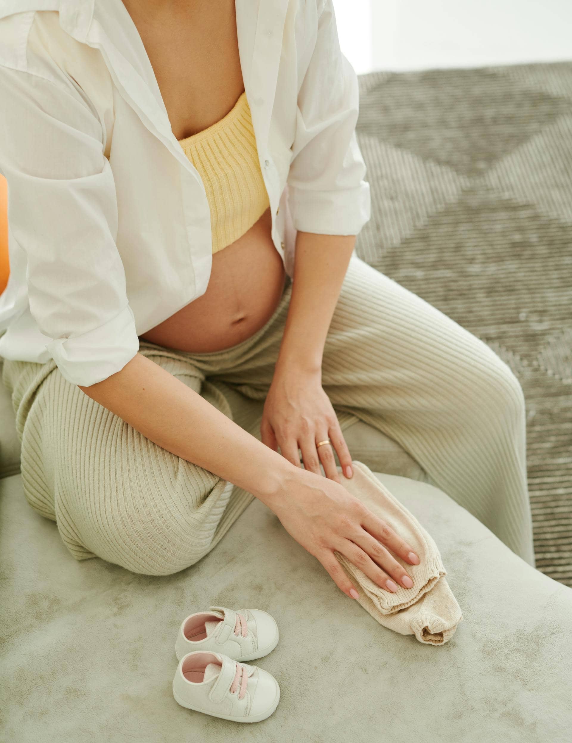 Pregnant woman during perimenopause folding baby clothes beside baby shoes.