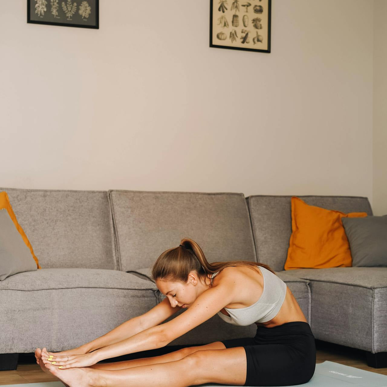 Woman stretching forward on a yoga mat in a living room, reaching for her toes during a seated stretch in front of a grey sofa.