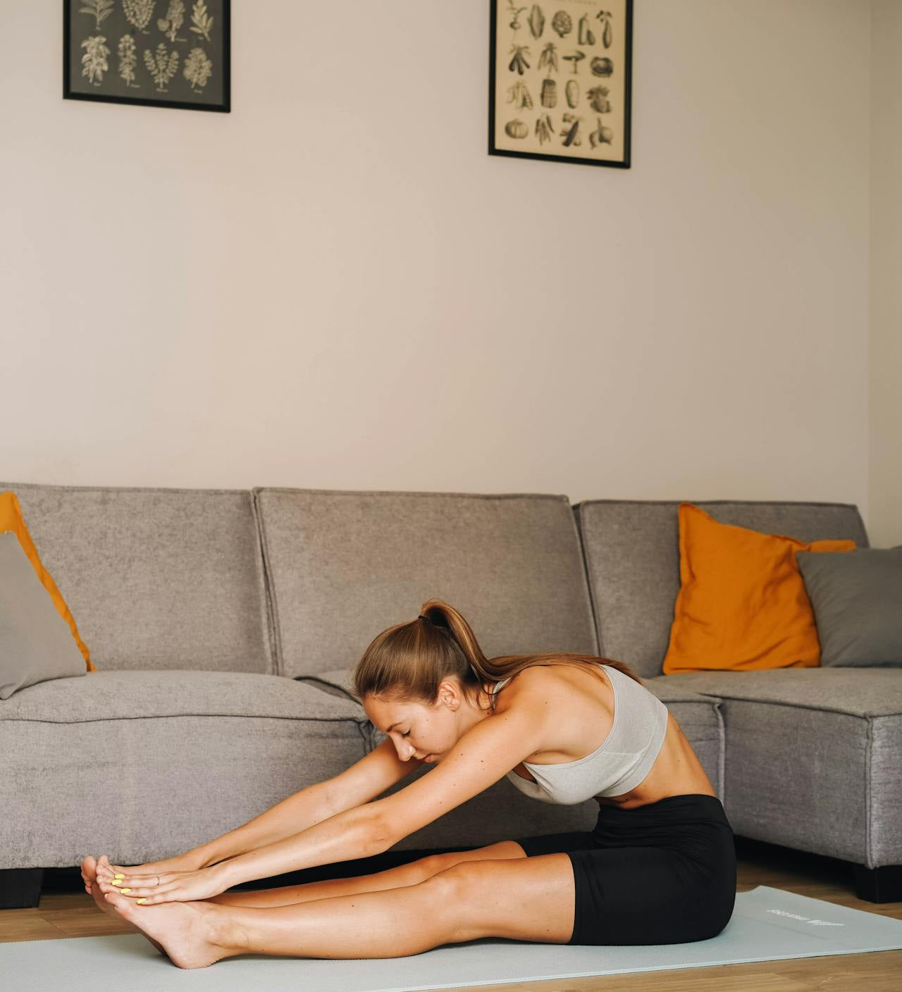 Woman stretching forward on a yoga mat in a living room, reaching for her toes during a seated stretch in front of a grey sofa.