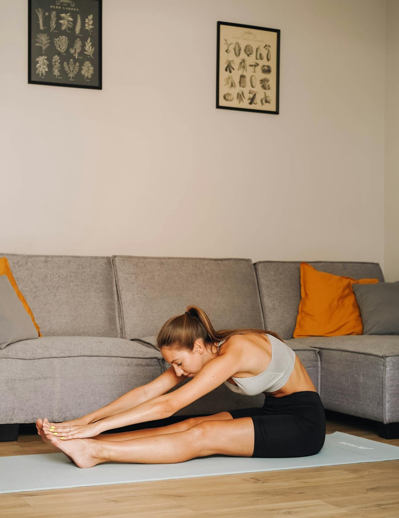 Woman stretching forward on a yoga mat in a living room, reaching for her toes during a seated stretch in front of a grey sofa.