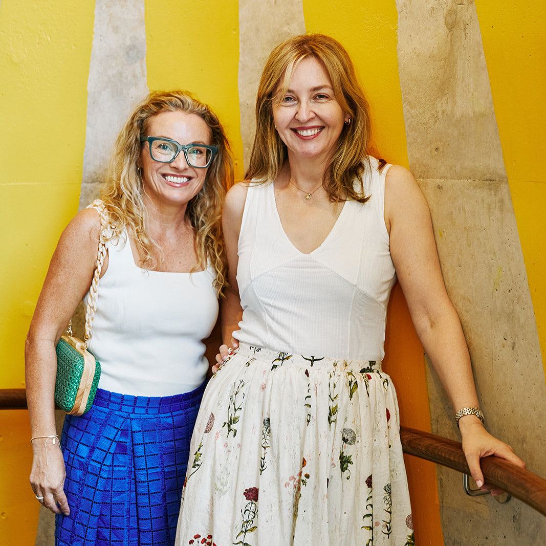 two women with blonde hair standing together as friends. Both in white tops and coloured skirts.